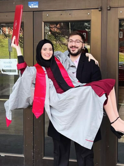 ally AboHana wearing a gray graduation gown with red accents and black heels, being carried by her husband while holding a red diploma cover aloft in front of glass doors.