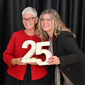 Nancy Swanger and Cheryl Oliver holding a large silver “25” sign, celebrating a 25-year milestone, in front of a black curtain backdrop.