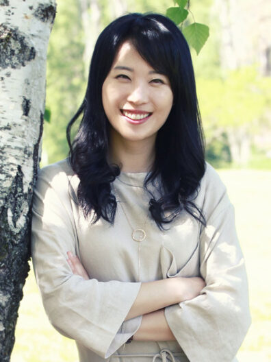Soobin Seo standing outdoors next to a birch tree, wearing a light beige long-sleeve dress with a thin belt and a circular pendant necklace, arms crossed. The background shows green foliage and sunlight.