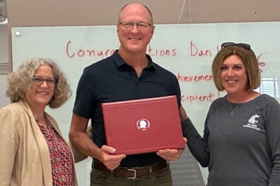 Dan Castles holding a red award folder while standing between Carson College Dean Deborah Compeau and Mariah Maki during a presentation of the Distinguished Alumni Award. A whiteboard with congratulatory text is visible in the background.