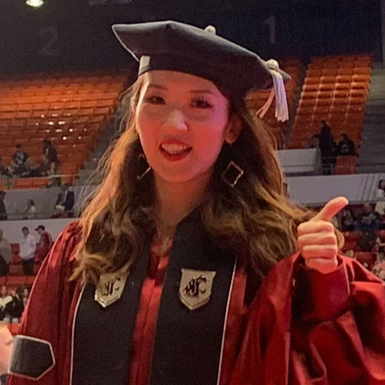 Yafang Li standing in graduation regalia at a large indoor ceremony, giving a thumbs‑up amid rows of seats and a crowd in the arena.