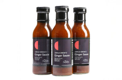 Three bottles of Unkle Drew’z Ginger Sauce arranged in a row, featuring dark labels with a red circle design on clear bottles filled with amber‑colored sauce.