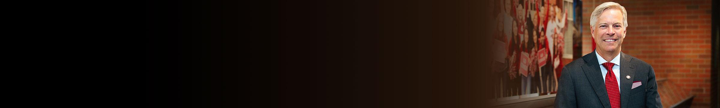 Carson College of Business Dean Chip Hunter standing in a suit with a red tie beside a wall display of people holding WSU signs, against a brick hallway backdrop.