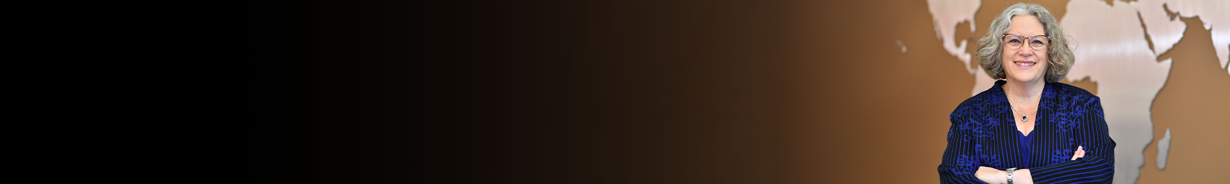 Carson College of Business Dean Debbie Compeau standing with arms crossed before a world map graphic on a tan background, positioned to the right of a wide black-to-tan gradient.