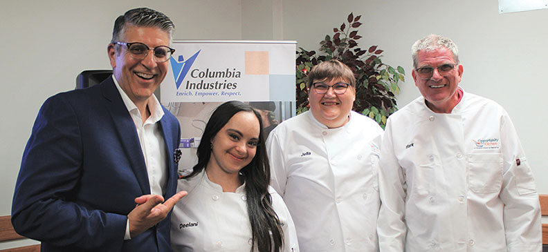 Michael Novakovich standing with Columbia Industries clients in white chef coats, posing together in front of a Columbia Industries banner and a potted plant.