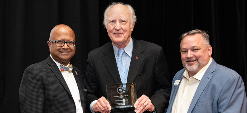 Terry Umbreit flanked by Carson faculty Dipra Jha and Anthony Anton, standing together as Umbreit holds an award against a dark backdrop.