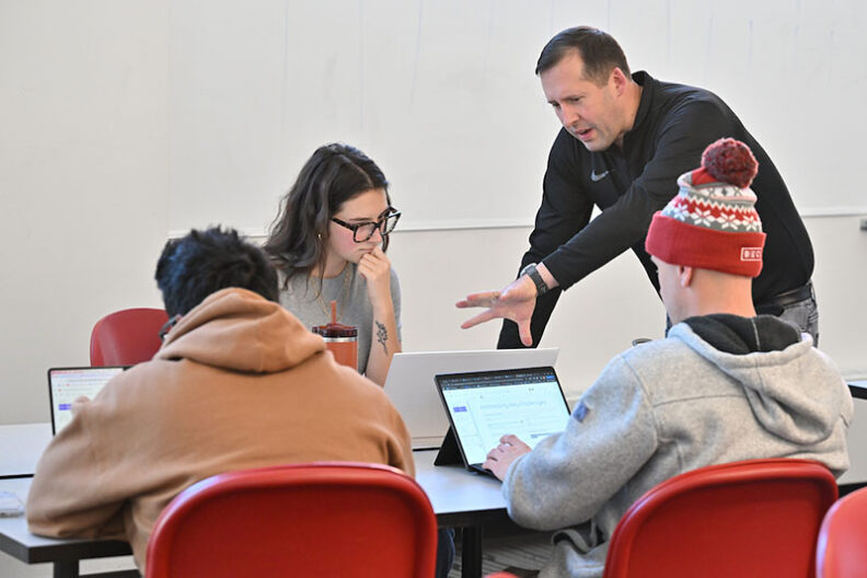 Professor Robert Crossler working with Eden Brooks and teammates seated at a table during the AI @ Carson Workshop, all focused on laptops in a classroom setting.