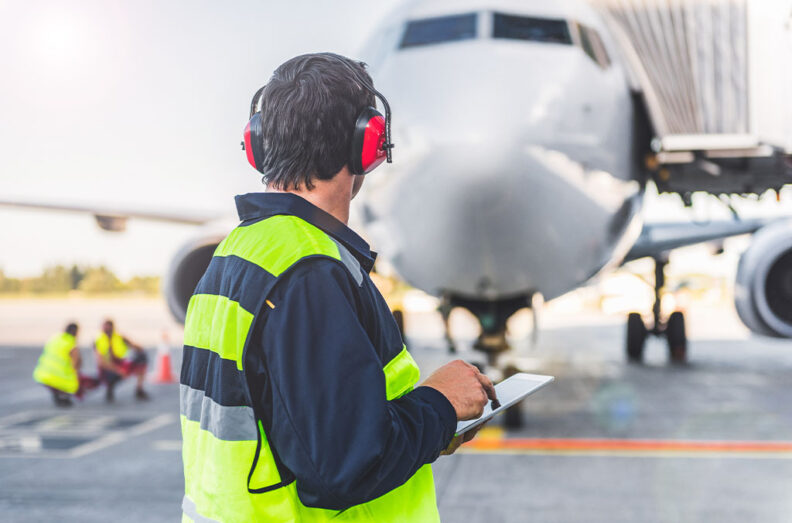 Airfield crew member in a high‑visibility vest and hearing protection holding a tablet while facing an airplane parked on the tarmac.
