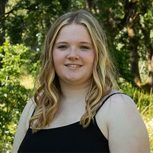 Lindsay Hayes standing outdoors in a sunlit wooded area, wearing a black sleeveless top with wavy hair resting over the shoulders.