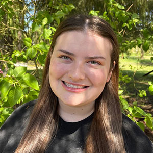 Zoie Rodman standing outdoors among bright green leaves, wearing a black top with long hair resting over the shoulders.