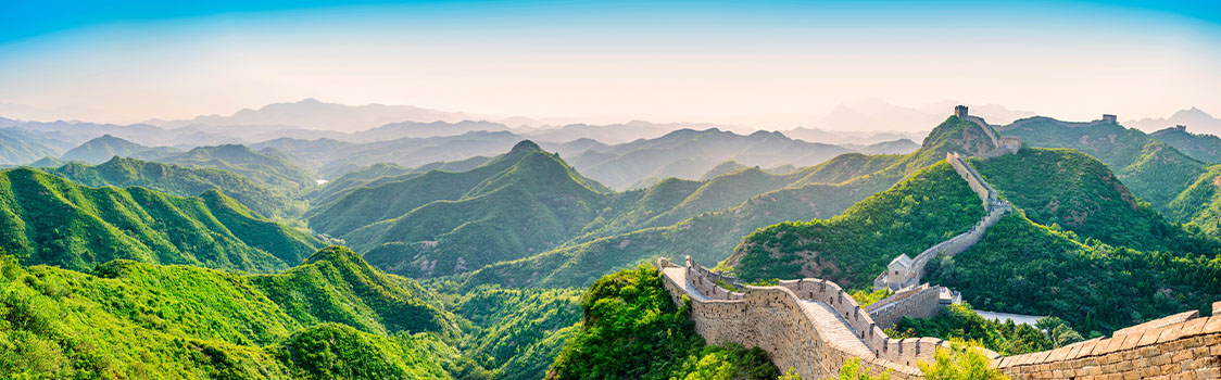 Panoramic view of the Great Wall of China winding over lush green mountains under a bright blue sky.