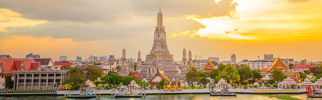 Wat Arun temple complex along the river in Bangkok at sunset, with golden light illuminating its central spire and surrounding rooftops.