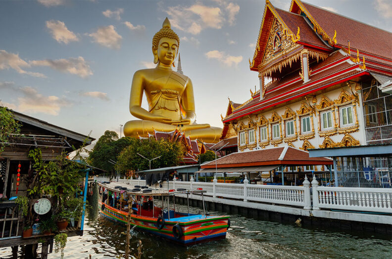 Large golden seated Buddha rising above an ornate temple with red and gold details beside a canal lined with colorful boats at sunset.