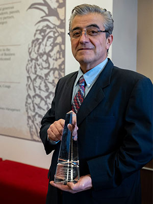Dogan Gursoy holding a tall, clear crystal award while wearing a suit and patterned tie, standing indoors near a wall with a large pineapple graphic.