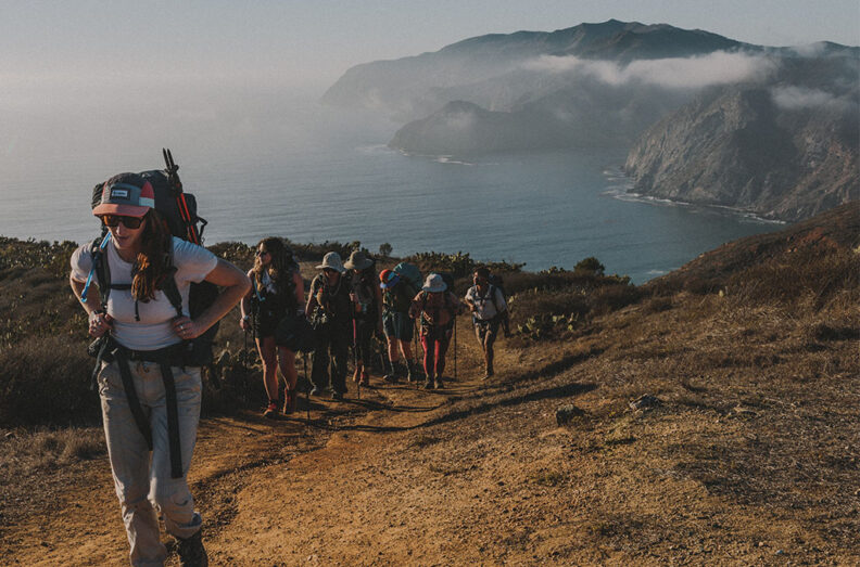 Group hiking along a coastal trail with backpacks, following a dirt path overlooking steep cliffs and the ocean below on a hazy day.
