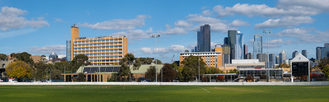 Wide view of a grassy sports field bordered by trees, with mid‑rise buildings in the foreground and a modern city skyline under a blue sky with scattered clouds.