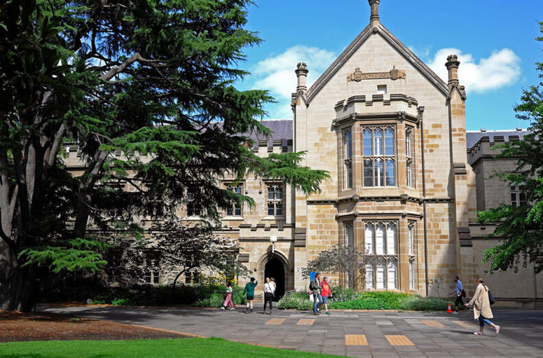 Historic stone university building with large arched windows and a central entrance, surrounded by trees and students walking across a paved courtyard.
