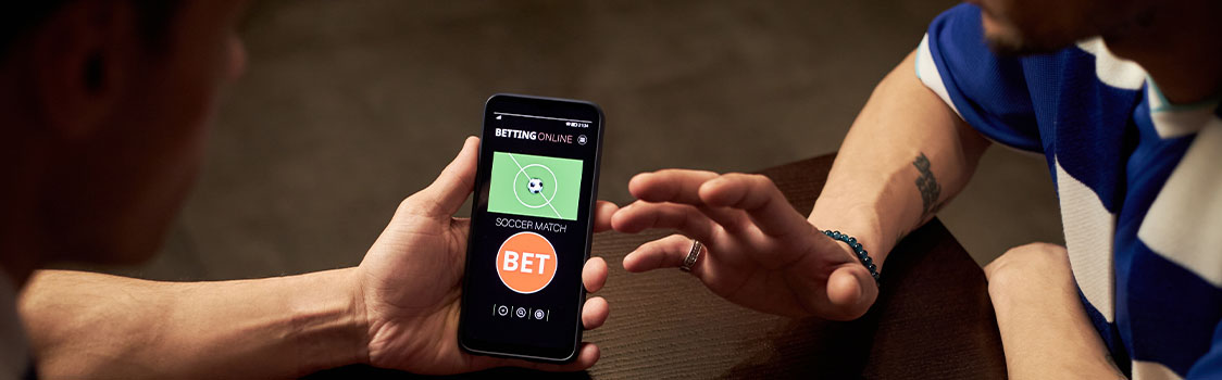 Two people seated at a table looking at a phone screen displaying an online soccer betting app with a prominent “BET” button.