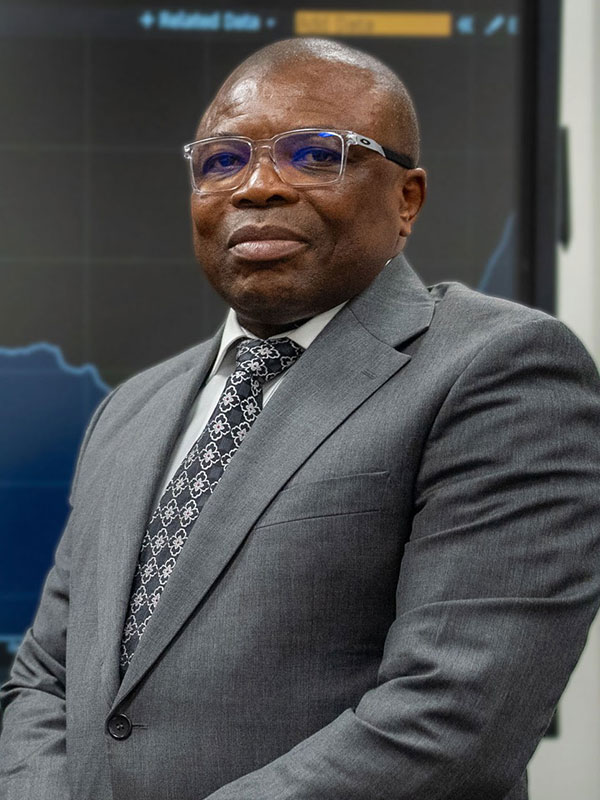 Kenneth Nwafor wearing a gray suit and patterned tie, standing in front of a large screen displaying financial charts and data.