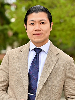 Long Nguyen wearing a tan blazer, light shirt, and patterned tie while standing outdoors with greenery in the background.