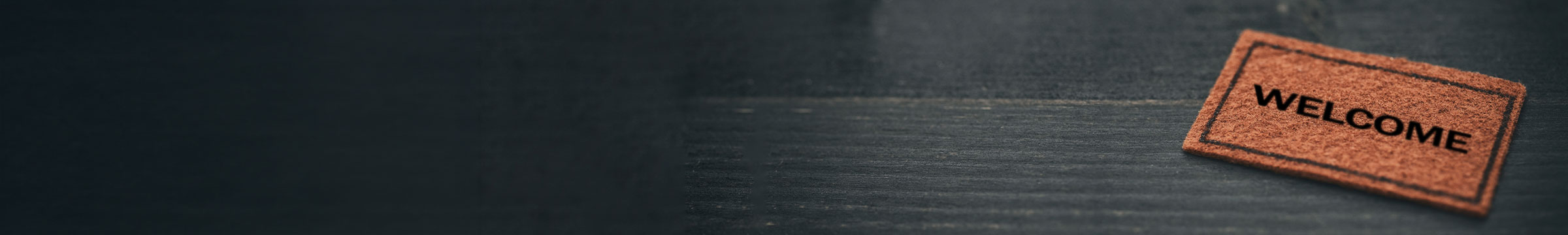 Small welcome mat with the word “WELCOME” on a dark wooden surface, positioned to the right with wide open space to the left.