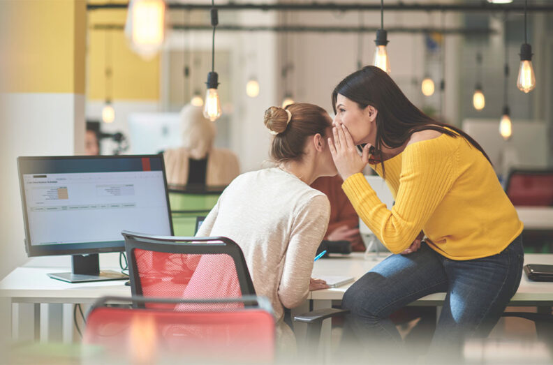 One person leaning close to another at an office desk, appearing to share a quiet comment while sitting beside a computer monitor in a modern workspace.