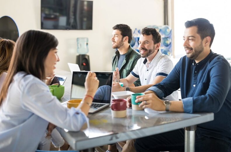 A group seated around a table in a bright workspace, talking over laptops, notebooks, and coffee mugs during a casual meeting.