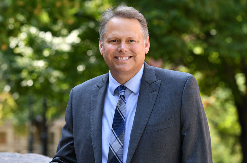 PhD Director Chuck Munson standing outdoors in a suit and tie, with sunlight filtering through green trees in the background.