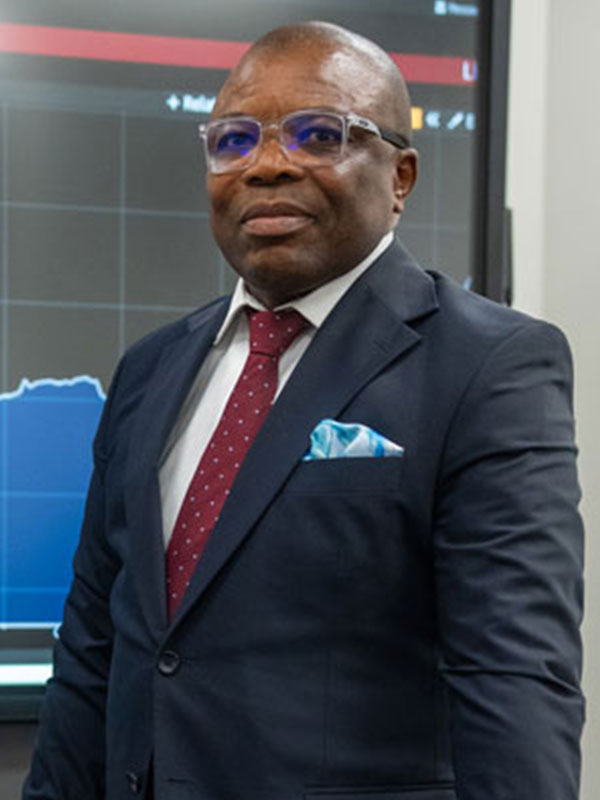Kenneth Nwafor wearing a dark suit and patterned tie, standing in front of a large screen displaying financial charts and data.
