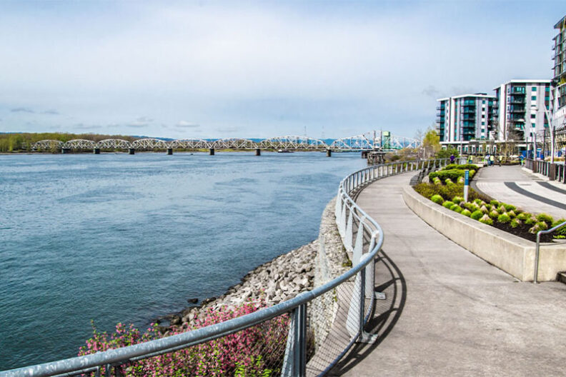 Waterfront path along the Columbia River in Vancouver, Washington, with modern riverfront buildings and the Interstate Bridge in the distance.