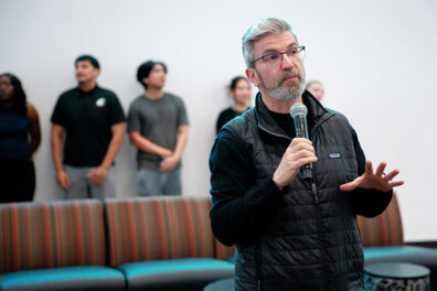 Chris Cooney speaks into a microphone while students prepare to present business ideas and mobile apps for a class project.