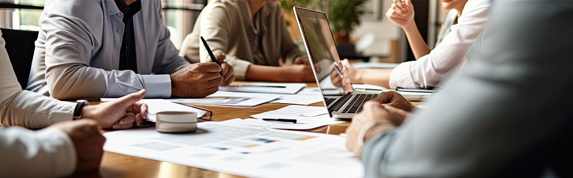 People gathered around a conference table review documents and a laptop during a collaborative business meeting.