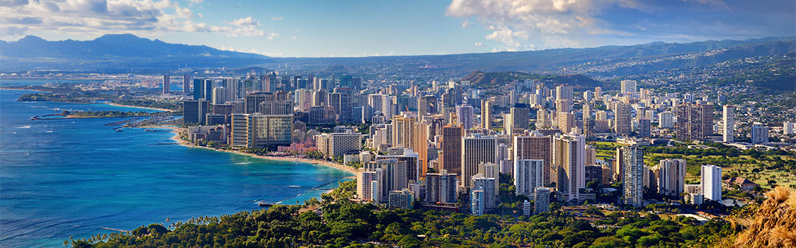 Panoramic view of Honolulu, Oahu, with dense high‑rise buildings along the coastline, turquoise ocean waters, and mountains in the distance.