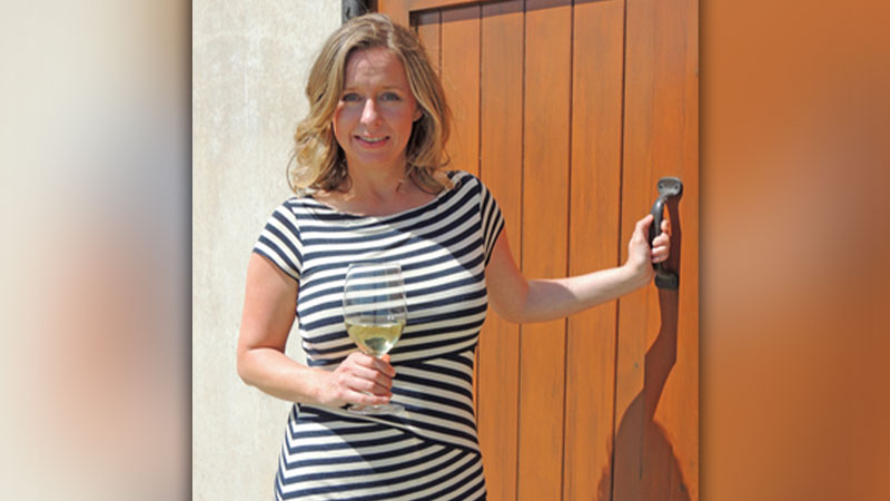 Jennifer Clapp in a striped dress holds a glass of white wine while opening a wooden door, standing in bright sunlight beside a stucco wall.