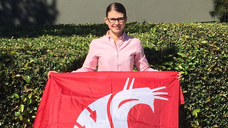 Kate (Stewart) Johnson standing outdoors holding a crimson Cougar flag, with hedges behind.
