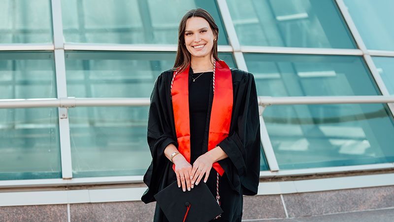 Meggie Kistler in a black cap and gown holding a diploma cover, with a red stole, standing outside a glass building.