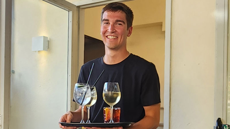 Noah Bridegwater wearing an apron holding a tray with wine glasses and drinks, standing in a doorway of an indoor space.