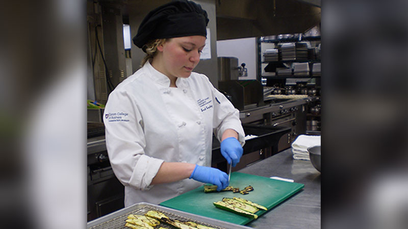 Sarah Beaudry in a chef’s jacket and gloves slicing grilled zucchini on a cutting board in a commercial kitchen.