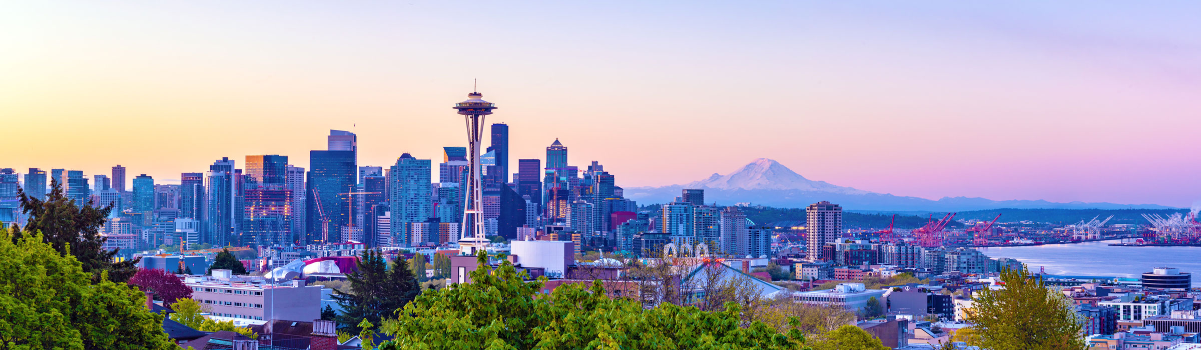 Panoramic Seattle skyline at sunset with the Space Needle centered among downtown buildings and Mount Rainier visible in the distance.