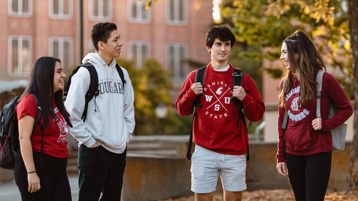 WSU students wearing Cougar gear and backpacks talk together outdoors on campus between classes.