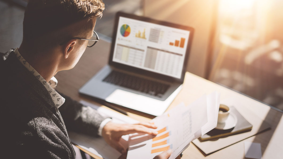 Person reviews financial reports beside a laptop displaying charts and spreadsheets in an office setting.
