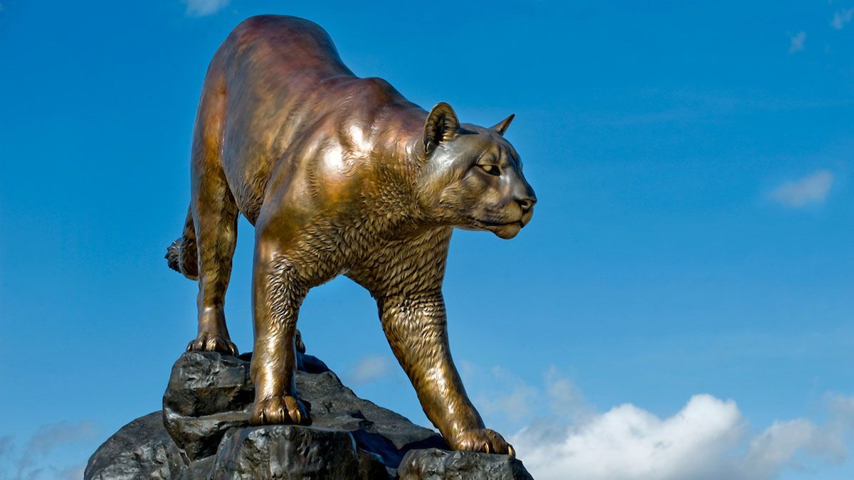Bronze cougar statue standing on a rock pedestal against a blue sky with scattered clouds.
