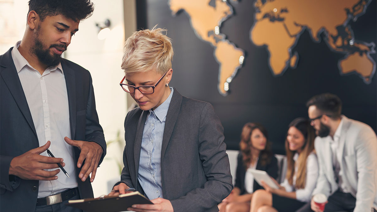 Professionals review information on a clipboard while colleagues meet in the background beneath a world map display.