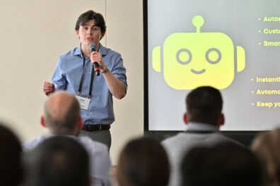Aidan Finnegan presents his venture during the BPC's final round, holding a microphone in front of a screen displaying a slide with a yellow robot icon and bullet points including 'Automated,' 'Customer,' 'Smart,' 'Instant,' and 'Automatic.'