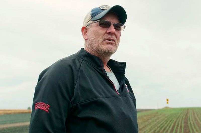 Douglas Poole standing in a field wearing a black jacket with red text on the sleeve and a light-colored cap, with rows of crops and an overcast sky in the background.