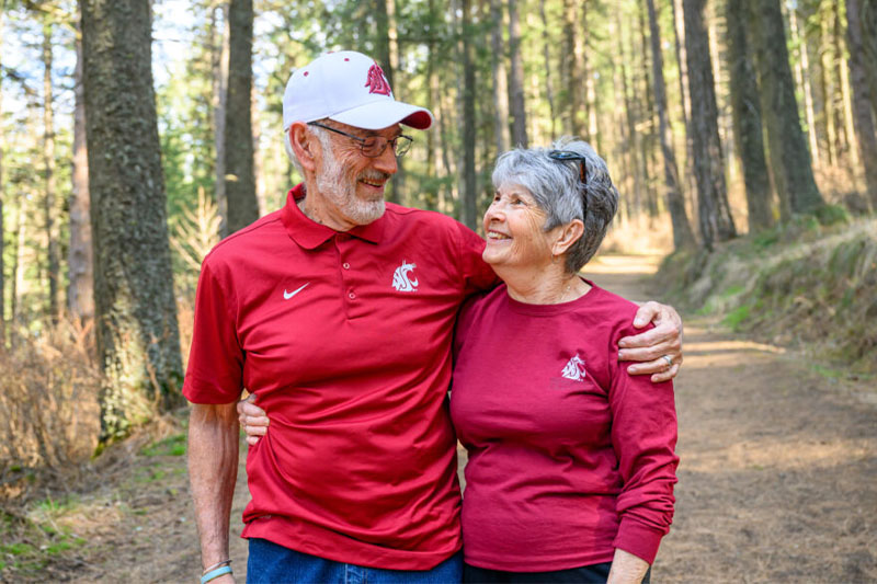 Two retirement-age individuals in red Washington State shirts with white cougar head logos standing on a forest path, with trees in the background.