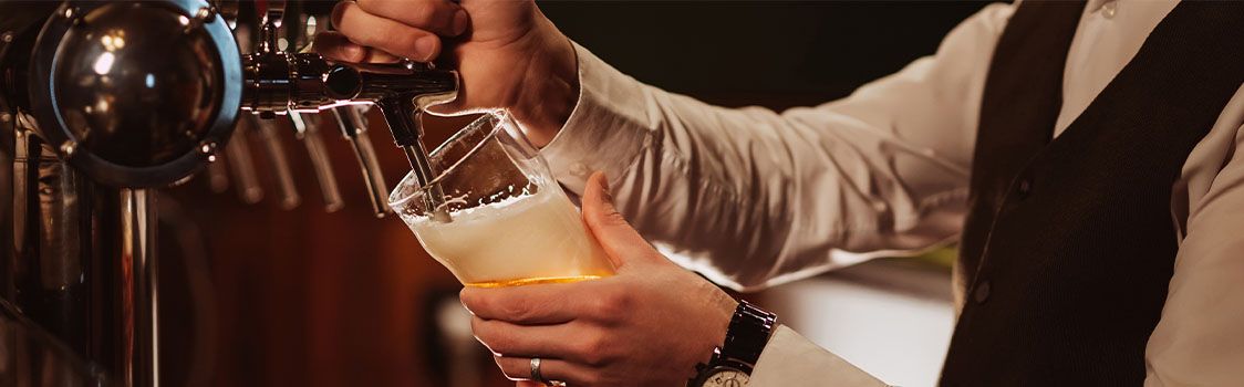 Person in a white shirt and dark vest pouring beer from a tap into a tilted glass, with foam forming at the top; bar setting with multiple taps in the background.