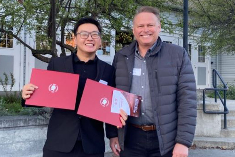 Yingwei (Wayne) Xu and PhD Program Director Chuck Munson standing outdoors holding red university folders.