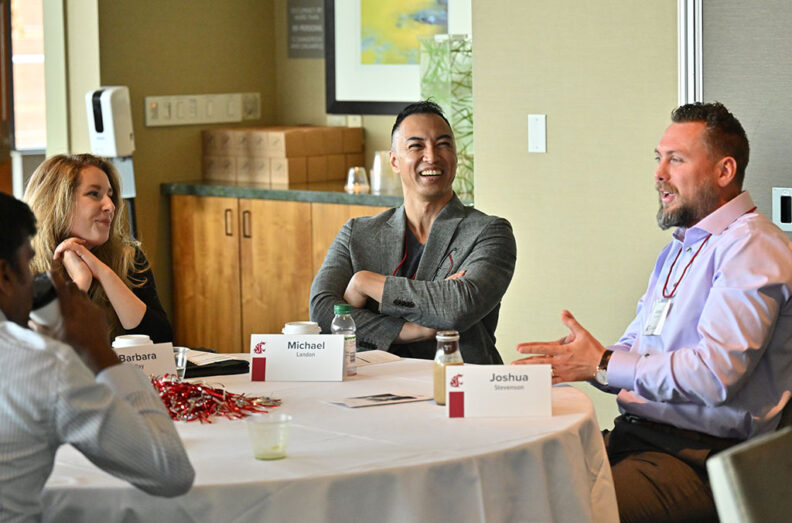 Group seated around a table at the EMBA Leadership conference with name tags, lanyards, and papers visible.