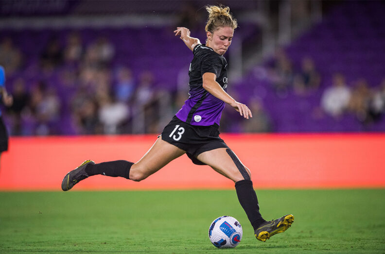 Celia Jimenez Delgado in a black and purple uniform, mid-stride about to kick a soccer ball.